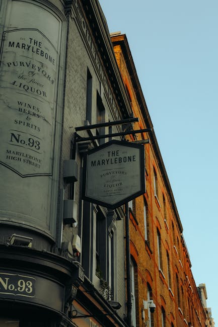 Photograph of the exterior of a historical building in Marylebone, showcasing a curved façade with a vintage-style black and white sign reading 'The Marylebone Purveyor of Fine Liquor, Wines & Spirits, No. 93, Marylebone High Street' mounted on the corner. The building features brickwork with large, rectangular windows, some with decorative white trim. The sign is attached with metal brackets and positioned above a street-facing window, under a clear blue sky. The building's façade appears well-maintained, highlighting its classic London architecture, with no visible dirt or damage, reflecting a neat and tidy appearance typical of a commercial establishment in the area. Marylebone Carpet Cleaning might use such buildings for their advertising or signage, emphasizing a clean and professional image associated with their domestic and commercial cleaning services.