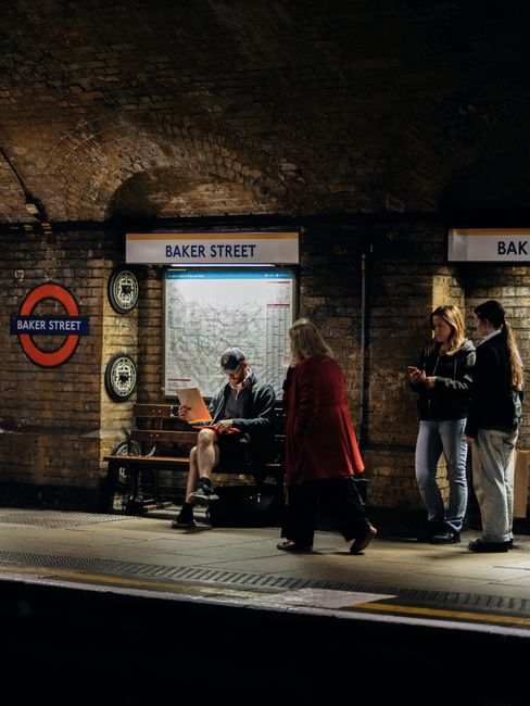 Indoor scene at a London tube station platform featuring a brick arch ceiling illuminated by warm lighting, with a sign displaying 'BAKER STREET'. There is a wooden bench where a man dressed in casual clothing and a cap is seated, reading a document or screen. Nearby, three women are standing; one woman in a red coat is facing away, while the other two, dressed in dark jackets and light jeans, are engaged in conversation, possibly using their phones. The platform surface appears clean with visible tactile paving for safety. The atmosphere suggests a busy urban environment, emphasizing the importance of maintaining cleanliness and hygiene standards within public transport facilities, as addressed by Marylebone Carpet Cleaning in relation to the standards for businesses like this location.