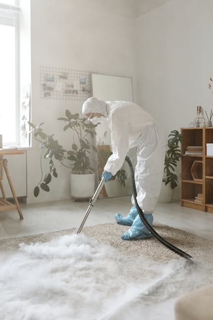 A person dressed in full protective cleaning gear, including a white suit, gloves, a face mask, and shoe covers, is kneeling on a beige carpet in a bright, modern living room. They are using a professional vacuum cleaner to perform deep cleaning and sanitisation of the carpet surface. The room features white walls, a large window allowing natural light, and a few potted plants, along with minimal furniture including a wooden sideboard and a small table. Visible cleaning tools and potted plants contribute to a tidy, hygienic environment, illustrating the high standards of cleaning maintenance promoted by Marylebone Carpet Cleaning for compliance with carpet hygiene standards in business settings.