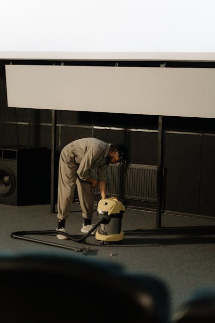 A professional cleaner dressed in a white protective suit, blue gloves, and shoe covers is performing deep cleaning on a beige shaggy carpet in a well-lit living room. The cleaner is using a vacuum or carpet cleaning machine with a hose attachment, focusing on removing dirt and stains from the surface. The room features white walls, a small wooden sideboard, potted plants, and a window allowing natural light to brighten the space. The scene depicts a thorough surface cleaning process aimed at maintaining hygiene and carpet freshness, consistent with domestic cleaning services offered by Marylebone Carpet Cleaning near Baker Street (W1).