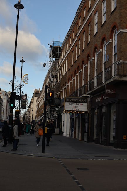 A busy street scene on Marylebone High Street in London showing a row of traditional brick buildings with large windows and balconies, some under construction with scaffolding visible. Pedestrians are crossing at a traffic light, with some carrying shopping bags. The street features classic street lamps and a variety of storefronts, including a sign for 'Everyman' theater and other shops. The sky is partly cloudy, casting natural light over the scene, highlighting the cleanliness and well-maintained appearance of the urban environment. This image represents a typical, well-kept commercial and residential area in Marylebone, where surface cleaning and maintenance are essential for maintaining hygiene and aesthetic appeal, aligned with services provided by Marylebone Carpet Cleaning.