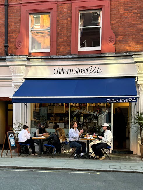 The exterior of Chiltern Street Deli in Marylebone features a traditional red-brick building with two white-framed windows on the upper floor. Below, a white facade houses the deli, with the name 'Chiltern Street Deli' elegantly inscribed above a deep blue awning that extends over the outdoor seating area. The outdoor seating includes five tables with black chairs, where patrons are enjoying their meals, some engaged in conversation, others appearing relaxed and content. The glass storefront reveals a glimpse of the interior, showing a cozy space with shelves and counter area. The sidewalk in front is clean and well-maintained, with a small chalkboard sign positioned to the left promoting the establishment. The overall scene is well-lit with natural daylight, contributing to a warm, inviting atmosphere, representative of a lively but tidy commercial environment in the Marylebone area, ideally maintained and suitable for a post-cleaning appearance. Marylebone Carpet Cleaning services ensure such spaces remain hygienic, bright, and welcoming for customers.