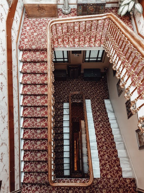 An aerial view of a historic staircase with ornate wooden banisters and detailed balustrades in a residential building, featuring red and beige patterned carpet on the stairs and landing. The stairs transition from carpeted sections to white marble steps, with a richly patterned rug on the landing area. Natural light illuminates the space, highlighting the polished wood finish and intricate details. The surrounding walls are adorned with framed artworks, and potted plants are placed near the staircase. The image showcases a well-maintained, elegant interior environment, emphasizing cleanliness and classic design, consistent with the services offered by Marylebone Carpet Cleaning in the W1 area for surface cleaning and maintenance.