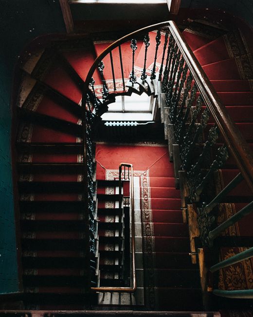 An overhead view of a spiral staircase inside a building, featuring dark wooden steps covered with a red carpet runner. The staircase has elegant black iron balustrades with decorative spindles, and a polished wooden handrail. Natural light filters in from a window at the bottom of the staircase, illuminating the area and highlighting the glossy, well-maintained surfaces. The surrounding floor is covered with a patterned rug, and the walls are painted in dark shades. This setting exemplifies a vintage or historic interior design, with an emphasis on surface and carpet cleaning to maintain hygiene and aesthetic appeal, as provided by Marylebone Carpet Cleaning.