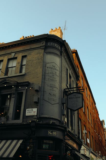 Exterior view of a historic building at the corner of a street in Marylebone, London, featuring a rounded facade with large windows and decorative architectural details at the top. The building is painted in muted tones with a sign displaying 'The Marylebone' and the address 'No. 98' below. There are street signs and a striped awning visible at ground level, with natural daylight illuminating the scene. The image emphasizes the urban architecture typical of the Marylebone area, pointing to the importance of professional cleaning and maintenance for historic and commercial properties, as highlighted by Marylebone Carpet Cleaning in their cleaning guide for homes near Regent's Park.