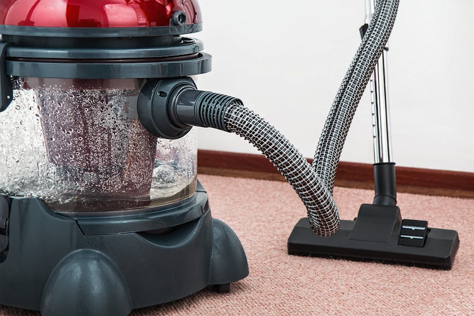 A close-up view of a vacuum cleaner with a transparent dustbin filled with debris, positioned on a pink carpet in a room with white walls and wooden skirting boards. The vacuum’s flexible hose, covered in a patterned fabric, is connected to a black floor attachment resting on the carpet. The image highlights the surface cleaning process, showcasing the equipment used for deep cleaning and sanitisation, and emphasizes the importance of maintaining clean and hygienic flooring surfaces in residential or commercial settings. The scene is well-lit, with the vacuum cleaner appearing to be in active use, aligning with the services offered by Marylebone Carpet Cleaning.