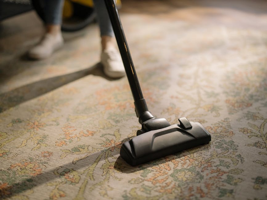 A close-up of a person's legs and feet wearing white sneakers and grey pants, operating a vacuum cleaner on a patterned area rug with floral motifs in beige, green, and orange tones. The vacuum cleaner's black head is pressed to the rug's surface, with the cord and handle extending upward. The room is well-lit with natural light, highlighting the clean, tidy appearance of the rug and the surface cleaning process. The setting appears to be a living room or similar residential space. The image emphasizes the deep cleaning and maintenance services provided by Marylebone Carpet Cleaning, as featured on their website page for same-day carpet cleaning quotes in W1U, MARYLEBONE.