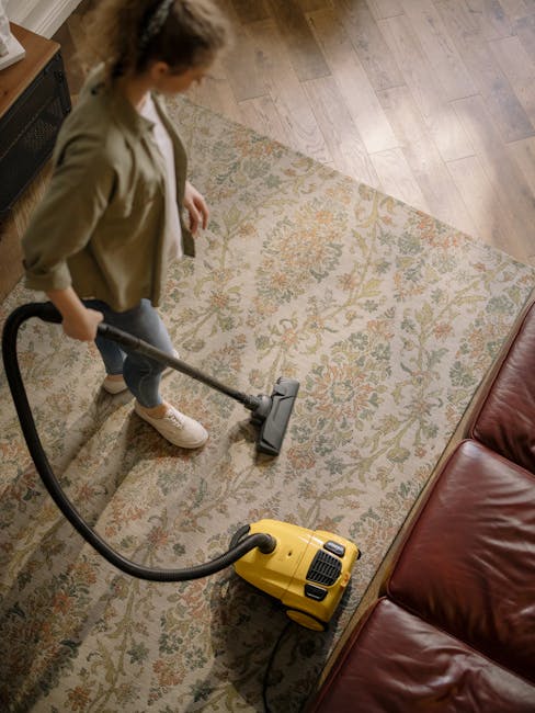 A woman using a yellow vacuum cleaner with a black hose attachment to conduct surface cleaning or deep cleaning on a decorative patterned area rug in a living room setting. The rug features intricate floral designs in beige, green, and orange tones, situated on a light wooden floor. A brown leather sofa is partially visible on the right side, and soft natural lighting illuminates the space. This scene exemplifies professional domestic cleaning services provided by Marylebone Carpet Cleaning to maintain hygiene and surface cleanliness.
