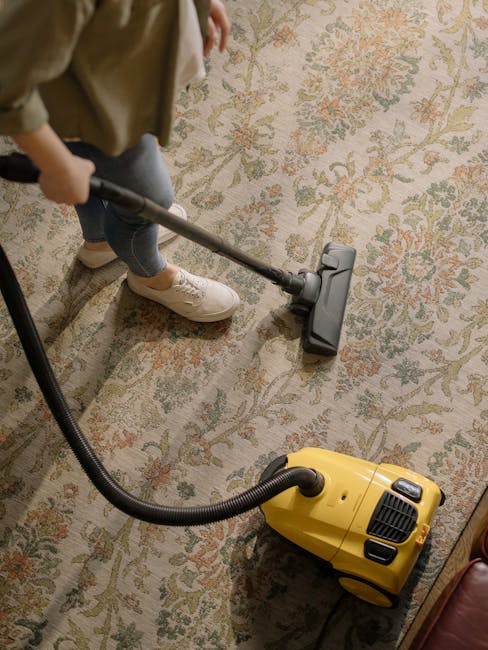 A close-up of a person's legs and feet wearing white sneakers and grey pants, operating a vacuum cleaner on a patterned area rug with floral motifs in beige, green, and orange tones. The vacuum cleaner's black head is pressed to the rug's surface, with the cord and handle extending upward. The room is well-lit with natural light, highlighting the clean, tidy appearance of the rug and the surface cleaning process. The setting appears to be a living room or similar residential space. The image emphasizes the deep cleaning and maintenance services provided by Marylebone Carpet Cleaning, as featured on their website page for same-day carpet cleaning quotes in W1U, MARYLEBONE.