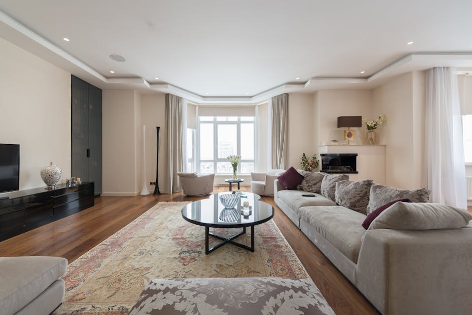 A bright, modern living room with white walls and a large bay window allowing natural light to fill the space. The room features a light-colored, plush fabric sofa decorated with mauve and grey cushions, positioned in front of a black fireplace with a small shelf. Adjacent to the window, there are two armchairs, one with a round small table between them. A dark wooden media unit sits along the wall, topped with decorative items including a ceramic vase. The room has polished wooden flooring, covered partly by an ornate rug. Visible furniture and surfaces appear clean and well-maintained, with no dust or clutter, emphasizing thorough domestic cleaning. The lighting is soft with recessed ceiling lights, enhancing the room’s inviting atmosphere, and the overall impression reflects a neat and hygienic living area as part of comprehensive surface cleaning and maintenance, fitting for property management in Marylebone flats. Marylebone Carpet Cleaning offers professional deep cleaning services to support landlords in maintaining mould-free carpets and hygiene in rental properties.