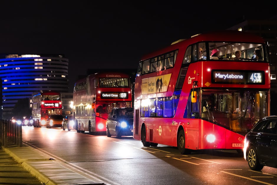 Nighttime street scene featuring three double-decker buses in red, with the front bus displaying urgent travel routes to Marylebone and Oxford Circus, parked along a city road illuminated by streetlights. The scene includes nearby black cars, modern office buildings with illuminated windows in the background, and a dark sky overhead. The buses are clean and well-maintained, with clear signage and lit interior spaces visible through the windows, conveying a sense of active urban transportation. The overall setting emphasizes the bustling nightlife and transportation hub, with professional lighting highlighting the shiny exteriors of the vehicles. This image showcases the importance of surface cleaning and maintenance of public transport vehicles in urban environments, relevant to the services provided by Marylebone Carpet Cleaning, as referenced on the webpage about permits for carpet cleaning at Marylebone events.