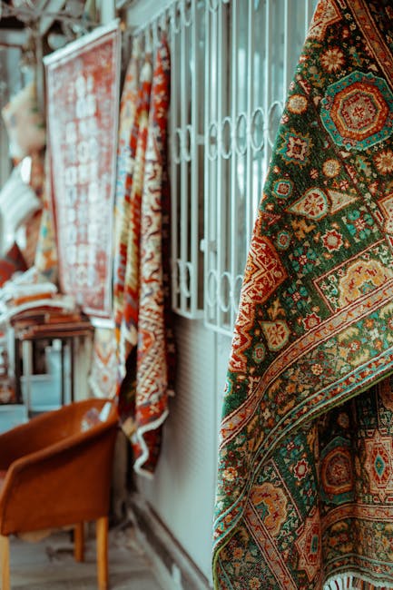 The image showcases a display of traditional Persian-style rugs hanging inside a well-lit room with natural sunlight. The rugs feature intricate patterns with rich colors, including reds, blues, and golds, and are draped over a metal railing or partition, revealing the detailed weaving craftsmanship. In the background, a wooden chair and a table with folded textiles are visible, suggesting a domestic or commercial setting focused on surface cleaning or rug care. The walls are light-colored, and the overall scene emphasizes cleanliness and attention to detail, aligning with the expertise of Marylebone Carpet Cleaning in deep cleaning and sanitization of woven surfaces, especially as part of Portman Square rug care. This environment highlights the importance of proper maintenance to preserve the beauty and hygiene of heritage and high-quality rugs.