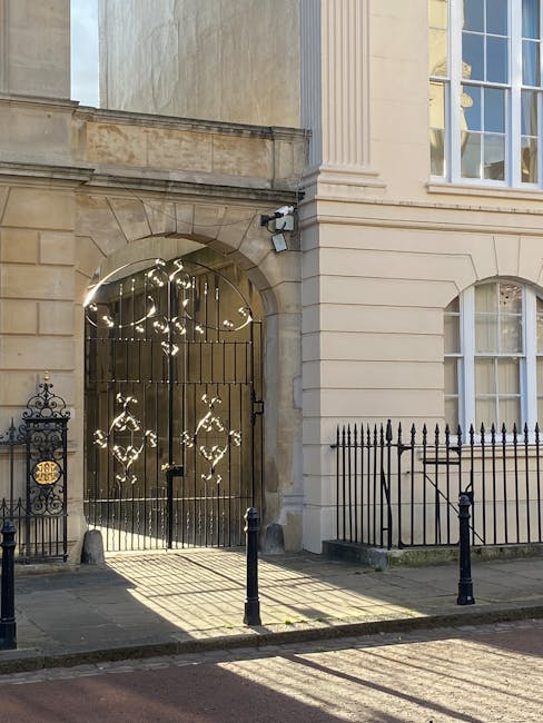 Photograph of an elegant exterior entrance to a historic building in Marylebone, featuring a black wrought iron gate with intricate decorative patterns and vertical bars, set within a stone archway. The building’s façade showcases light-colored, smooth stonework with classic architectural detailing, including large windows with white frames and decorative molding. Sunlight casts distinct shadows on the pavement and the building, highlighting its clean, well-maintained appearance. A security camera is mounted above the gate, and black metal street bollards line the sidewalk to protect pedestrians. The overall scene reflects the refined urban setting of Marylebone, emphasizing the building's pristine condition and the neat, tidy environment that Marylebone Carpet Cleaning aims to maintain through professional surface cleaning and deep cleaning services to eliminate smoke and odour residues from carpets, ensuring hygienic, fresh interiors.