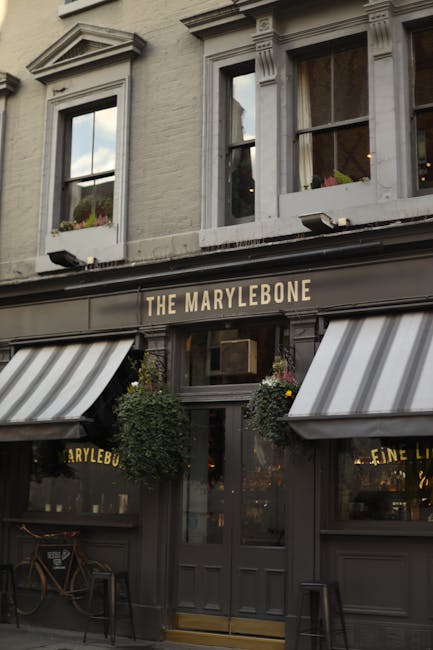 Exterior view of The Marylebone pub showing its grey-painted facade with decorative architectural molding and two large windows on the upper level. The entrance features a black sign with gold lettering reading 'The Marylebone', flanked by black and white striped fabric awnings extending over the sidewalk. Potted plants with hanging greenery and flowers decorate either side of the doorway. The street scene captures a typical London pub ambiance with outdoor seating visible inside through the glass. Marylebone Carpet Cleaning logo and service details are not visible in this image, as it focuses on the pub's exterior architecture and entrance setup.