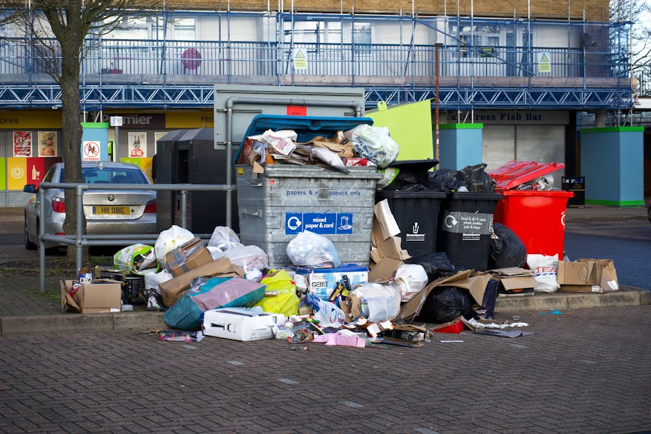 An outdoor street scene showing a large pile of mixed residential and commercial waste, including cardboard boxes, plastic bags, paper, and discarded packaging, overflowing from multiple waste bins and containers. The waste is spilling onto the cobblestone pavement near a parking area with a silver car, situated in front of a building with storefronts, including a seafood shop and fish bar. The area is lit with natural daylight, and the waste appears untidy and uncontained, highlighting the importance of proper waste disposal practices in compliance with Westminster Council regulations for carpet waste and disposal. Marylebone Carpet Cleaning emphasizes the significance of proper cleaning and hygiene standards in maintaining community cleanliness.