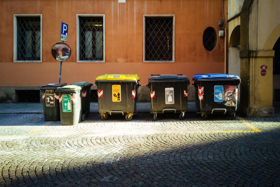 An outdoor street scene showing a large pile of mixed residential and commercial waste, including cardboard boxes, plastic bags, paper, and discarded packaging, overflowing from multiple waste bins and containers. The waste is spilling onto the cobblestone pavement near a parking area with a silver car, situated in front of a building with storefronts, including a seafood shop and fish bar. The area is lit with natural daylight, and the waste appears untidy and uncontained, highlighting the importance of proper waste disposal practices in compliance with Westminster Council regulations for carpet waste and disposal. Marylebone Carpet Cleaning emphasizes the significance of proper cleaning and hygiene standards in maintaining community cleanliness.